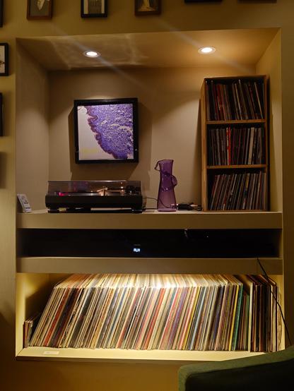 A photo of a record player on a shelf. On the wall above hangs a picture frame you can hang the record sleeve in that's now playing. To the right are three shelves, stacked, full of 7 inch records. Below all of this is one more shelf which houses over 100 LPs.