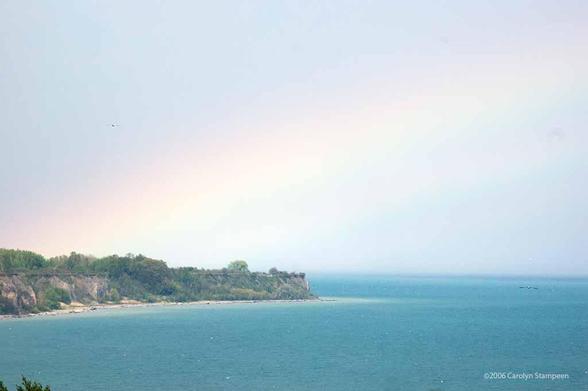Cliffs are rising from a narrow sandy beach, covered with trees, to make a point of land with water between us and it.  The water is blue, the sky before us has the remnants of the drizzly day while the sun breaking out of the clouds behind us has created a rainbow  that arches from above the land to the left to the top right of the image.