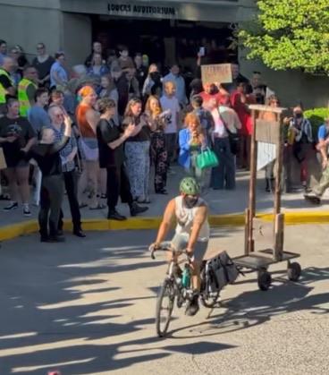 À person in à green cycling helmet cycles with a new style of trailer wagon in the form of a French decapitation device dating to the 17th and 18th centuries. The crowd clap and cheer.