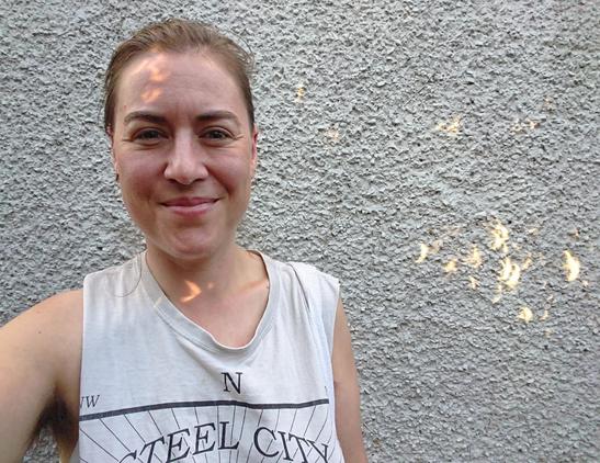 A white person smiling with crescent-shaped glints of light on their neck and forehead. They're standing in front of a grey wall with more crescent-shaped glints of light. (These are projections of the solar eclipse, because the light was passing through the gaps between leaves on a bush just behind the camera)