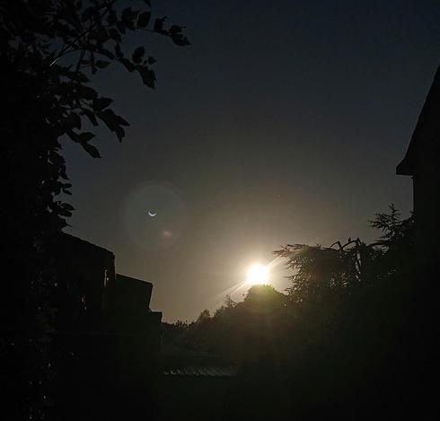 A very darkened photo of the sun looking normal, but a lens flare that's a very skinny crescent shape. In the foreground, some silhouettes of bushes and houses.