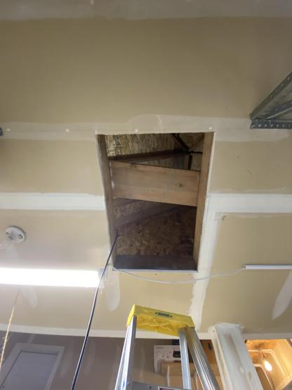 a rectangular hole in a semi-finished ceiling of a semi-finished room, a column and the top of a ladder also visible, and a bare-bulb light. through the hole is a crossing plank of wood and some chipboard, angled