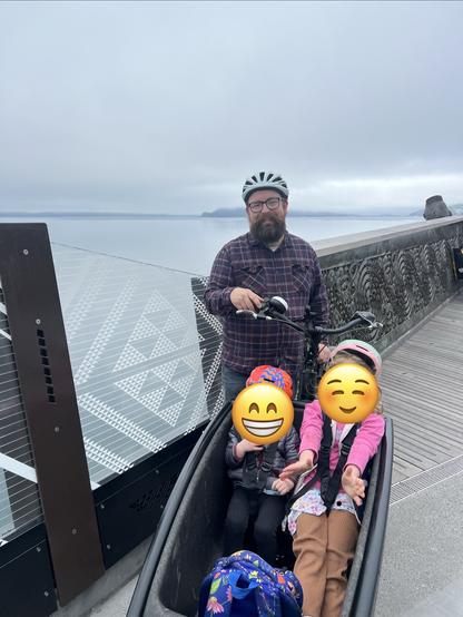 I stand, wearing a helmet standing next to a cargo bicycle with two children in the front. The kids have their faces redacted with emoji. The backdrop features a calm body of water and a cloudy sky.