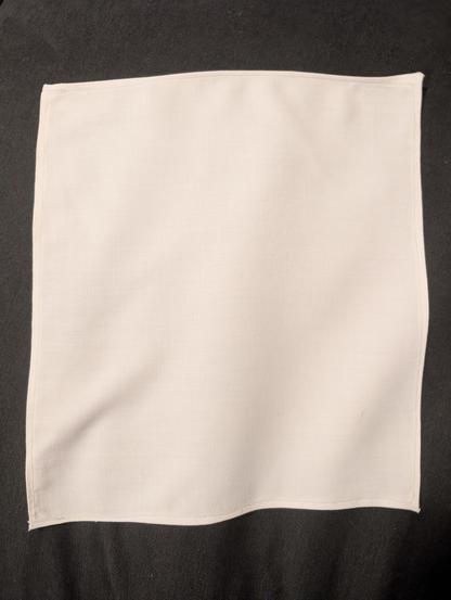 A white pocket square with straight stitching on the edges that are mostly straight.  The fabric has been recently ironed but has slight rippling.  It is resting on a dark grey couch cushion to provide nice contrast.