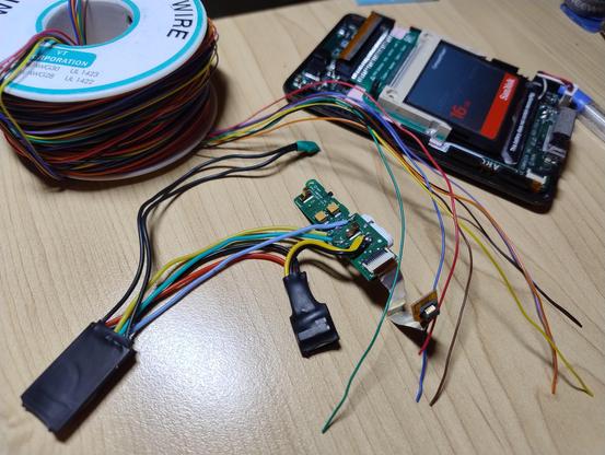 Half an opened iPod on a desk with a compact flash card inside it, and the headphone connector board next to it with a couple of other devices in heat shrink wrap wired onto it. A spool of colourful, thin wires in the background