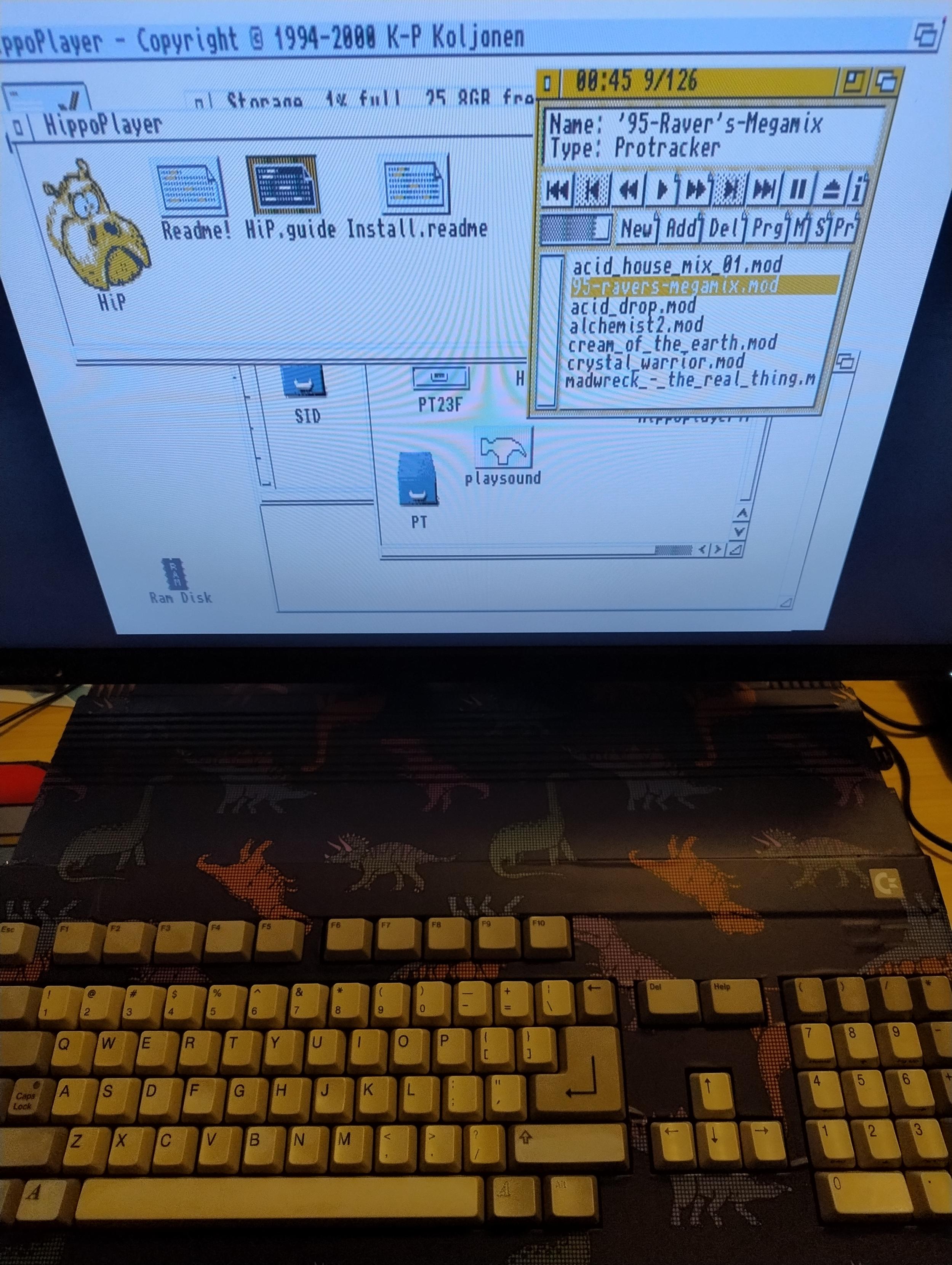 An Amiga 500 with a pixel dinosaur wrap in front of a monitor that's showing the Amiga desktop and HippoPlayer playing, yes, a track named '95 Raver's Megamix. 