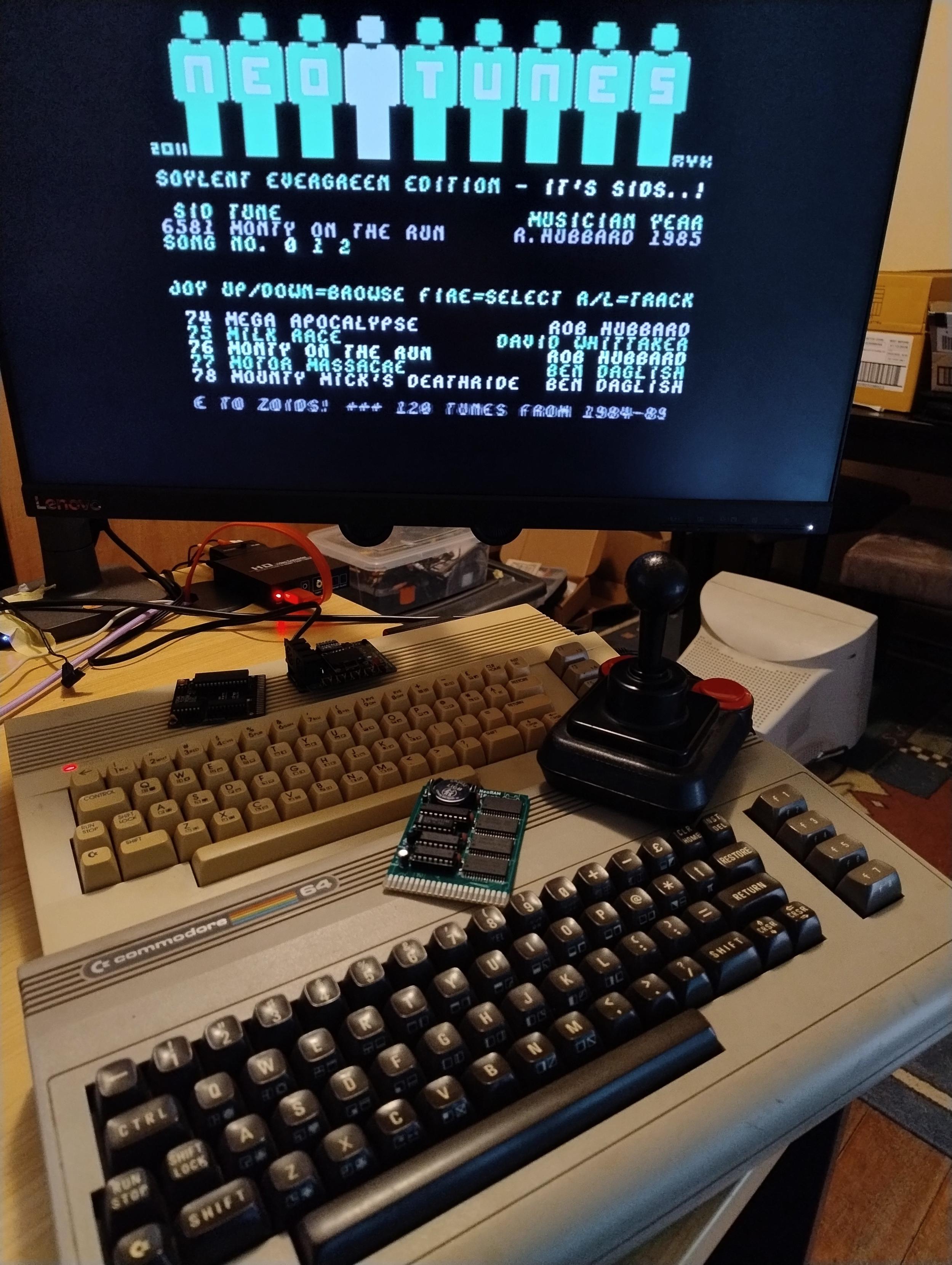 Two Commodore 64s, a brown breadbin and a while 64C style one on a desk. A joystick and a few cartridge boards are on top, and a monitor above them is showing the display for NeoTunes, a program to play SID music from a large collection.