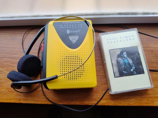 A bright yellow portable cassette player with some basic retro style headphones next to a copy of Graceland by Paul Simon