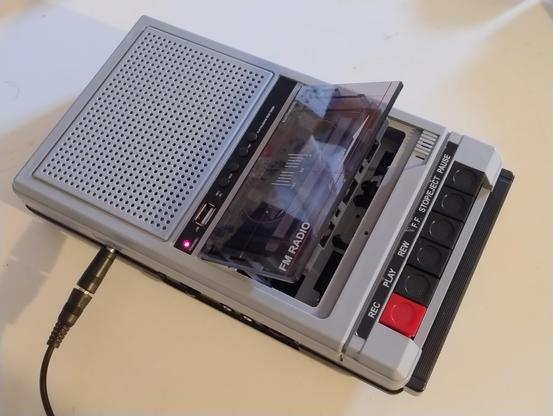 An old school looking grey cassette player with a handle and big speaker, but also with a very prominent USB port on top.