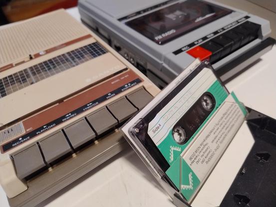 A 1980s Sanyo DR101 tape player next to a modern equivalent with USB input and output, and an old audio cassette with a hand written label reading 