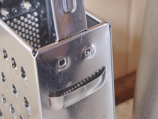 Close up on the side of a kitchen vegetable or cheese grater at an angle so it looks like one slicing blade is a mouth below two eyes and the holes on the back framed so they look like teeth in the mouth