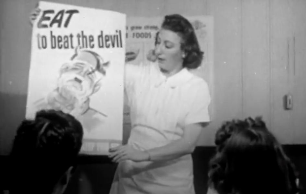 Black and white frame from an old movie with a woman holding up a poster in front of some people looking on. The poster reads 