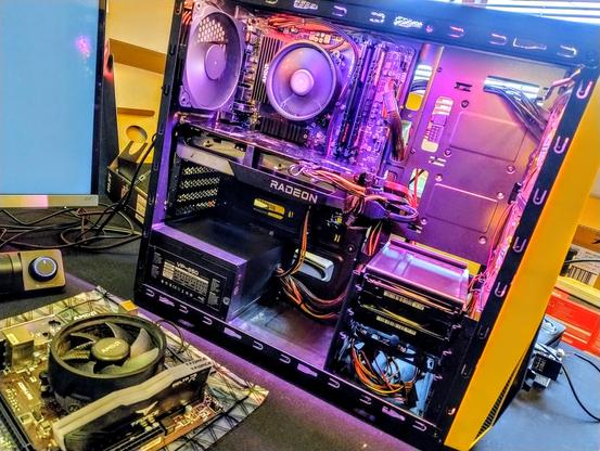 Inside of a PC on a table with another motherboard next to it. The inside of the case is lit up with bright LEDs, mostly showing purple tones