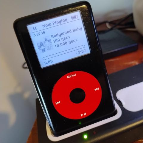 A U2 Special Edition iPod in a charging dock, the monochrome screen has now playing info up for Hollywood Baby by 100 gecs