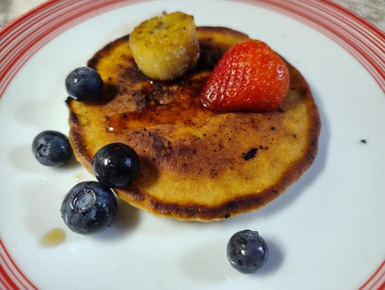 A small pancake on a plate with some blueberries, half a strawberry, a slice of fried banana, and a light drizzle of maple syrup 