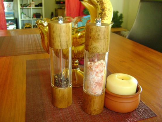 Transparent salt and pepper grinders on a table in front of an amber glass bowl with a couple of bananas in it.