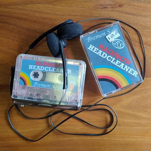 A transparent audio cassette player with a copy of a tape called Rainbow Headcleaner by Bremen Saki in it, next to a tape case for the album that has a price sticker on it marked at $420.69, the whole thing surrounded by some really cheap looking black headphones
