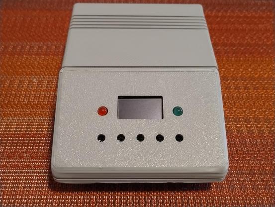 A small grey box with an angled front with a small screen, red and green LEDs and spots for 5 buttons