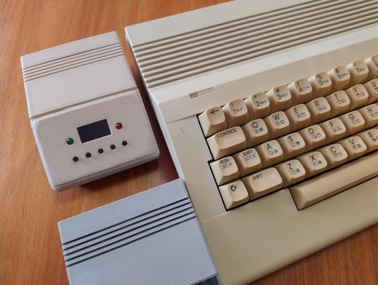 A small printed case with buttons and a screen next to a Commodore 64 showing an aesthetic similarity and colour. There's part of a grey version of the same case visible in the foreground.
