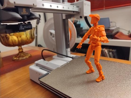 An orange 3d printed posable figure holding a spanner standing on the bed of a 3d printer, looking up at the print head