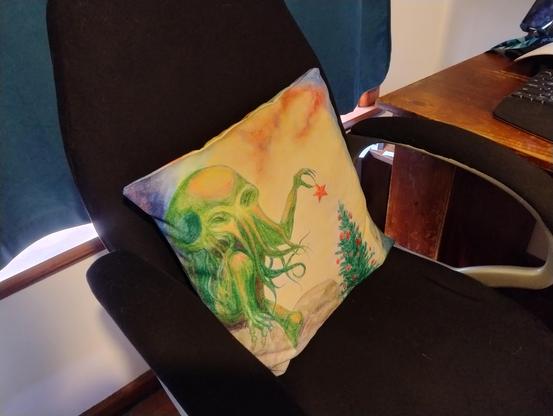 An office chair with a cushion on it, printed with a picture of Cthulhu delicately placing a star on top of a Christmas tree.