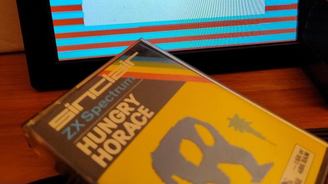 Close up of the cassette case for Hungry Horace for the ZX Spectrum, in front of a small screen with red and blue stripes in the border 