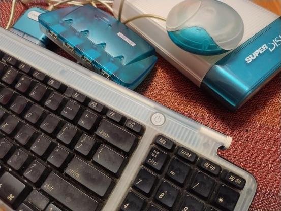 A keyboard, mouse, floppy drive, and USB hub all in the blue and white transparent plastic aesthetic of Apple computers of the late G3 era. 