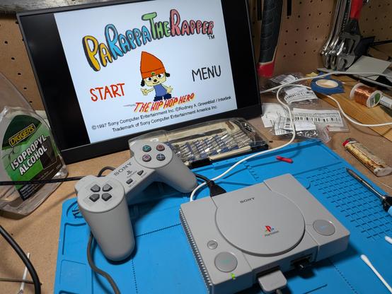 A PlayStation Classic with a small USB drive in port 2, on a work bench hooked up to a monitor showing the start screen for Parappa the Rapper 