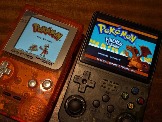 A Game Boy Pocket in a transparent orange case displaying the Pokemon Red start screen, next to a new handheld console showing the Pokemon FireRed version 

